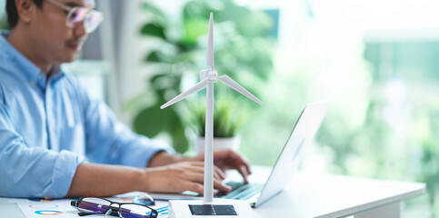 A man is typing on a laptop in front of a wind turbine model. Concept of innovation and progress, as the man works on a project related to renewable energy