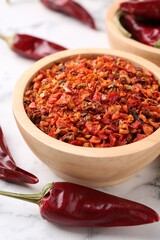 Chili pepper flakes in bowl and pods on white marble table, closeup