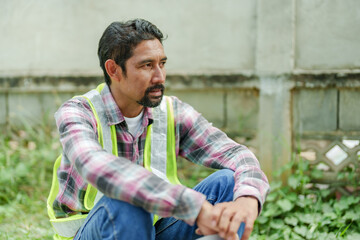 Asian male construction worker sitting at building site, wearing reflective vest and holding hard...