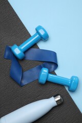 Two dumbbells, yoga mat, fitness elastic band and thermo bottle on light blue background, flat lay