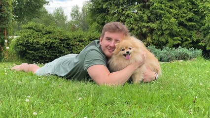 Young man is lying on green grass in park with his cute little Pomeranian Spits dog, guy play, hug...