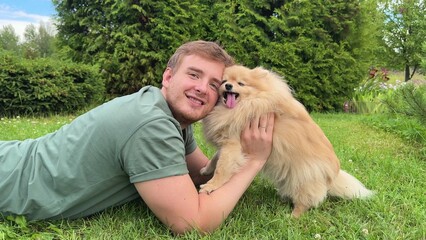 Young man is lying on green grass in park with his cute little Pomeranian Spits dog, guy play, hug...