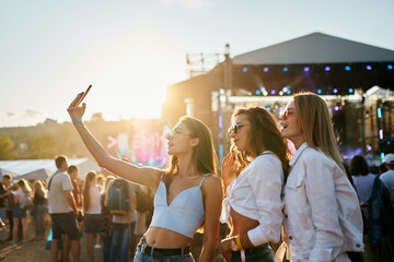 Girls in summer attire enjoy sunset at coastal music festival, stage lights glow in background. Youth share moments, dance to live performances, sandy beach venue vibrates with fest energy. - Powered by Adobe