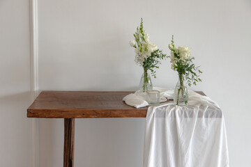 A rustic wooden table is elegantly decorated with two vases of white flowers and a flowing white...