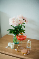 Bouquet of pink peonies in glass vase, candles, glass with pink cocktail, cosmetics, things for women, decorations on wooden table. Spring flowers.