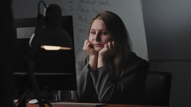A tired young blonde businesswoman sits at workplace at the table and looks at the computer monitor with a thoughtful, detached gaze, blinks and props chin with palms clenched into fists. The concept