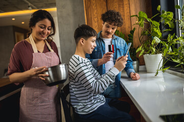 Latina mother cooking in the kitcher, stirring in the pot while her husband and son are ordering online on the tablet with credit card