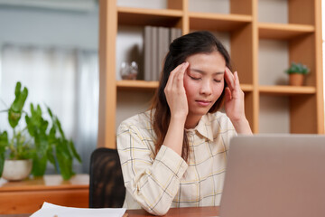 Asian woman presses hand to head, eyes closed, portraying either profound focus or onset of fatigue. Office worker rests head in hand, embodies momentary retreat from digital work demands.