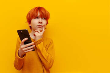 pensive red-haired child boy using smartphone and planning on yellow isolated background, puzzled teenager winner with orange hairstyle thinks and imagines and looks away