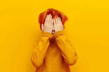 scared red-haired child boy hiding and covering his face with his hands on a yellow isolated background, excited teenager with orange hairstyle afraid and shy