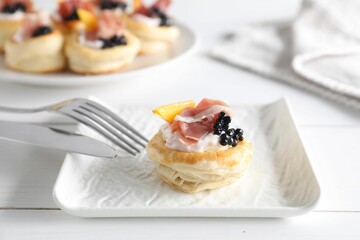 Delicious puff pastry with cream cheese, ham, black caviar, peach and cutlery on white wooden table, closeup