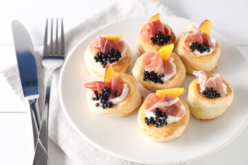 Delicious puff pastry with cream cheese, ham, black caviar, peach and cutlery on white wooden table
