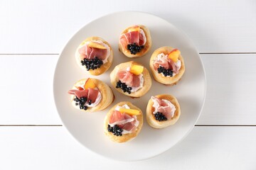 Delicious puff pastry with cream cheese, ham, black caviar and peach on white wooden table, top view