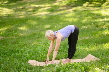 Fototapeta premium Beautiful middle aged woman practicing yoga in park. Ardha Uttanasana pose. Woman performs standing asana, half-bending with straight back, on mat on grass on summer. Health concept, yoga in nature.
