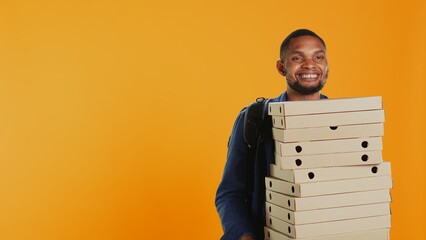 Male pizzeria courier carrying huge pile of pizza boxes in studio, preparing to deliver meal order...