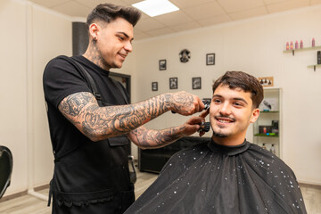   tattooed barber cutting the hair and beard of a young, dark-haired client.