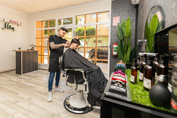   tattooed barber cutting the hair and beard of a young, dark-haired client.