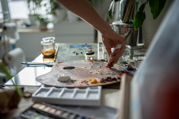 Hand of female artist squeezing oil, acrylic paint out of tube onto palette. Palm of talented creative woman preparing to start work on painting, organizing workplace. Closeup, table with art supplies
