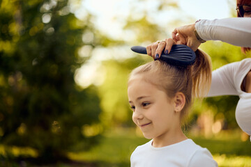 Smiling little girl getting her hair styled by her mom, at the park, using a comb.