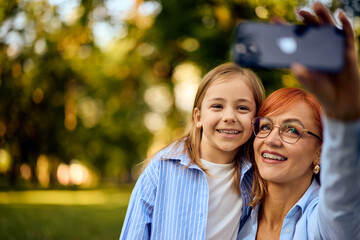 Fototapeta premium A smiling ginger-haired mom using a phone and taking photo of her and her daughter, at the park.