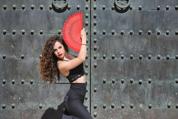 Portrait of young, beautiful, brunette woman with black top and skirt, dancing flamenco with a red fan with an old, black metal door in the background. Flamenco concept, dance, art, typical Spanish.