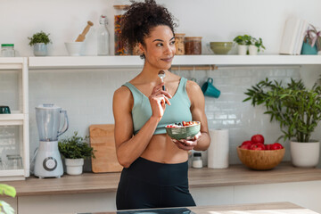 Athletic woman eating a healthy fruit bowl while looking forwards in the kitchen at home