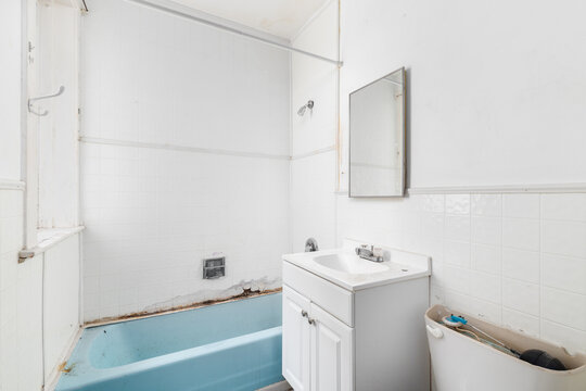 A bathroom with water damage and falling apart with white square tiles, a white bathroom cabinet, and a dated blue bathtub ready to be renovated.