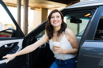 Woman smiling and holding a coffee cup as she steps out of her car on a sunny day, taking a...