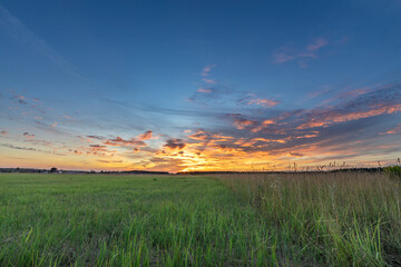 A picturesque view of a meadow at sunset, showcasing a vibrant sky with streaks of orange, pink,...