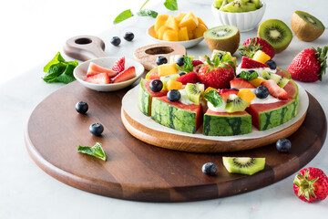 A homemade watermelon pizza surrounded by fresh fruit, next to a sunny window.