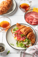Croissant sandwich with salami sausage, cheese and cucumber in a plate with cup of tea for tasty breakfast on white tile table
