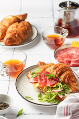 Croissant sandwich with salami sausage, cheese and cucumber in a plate with cup of tea for tasty breakfast on white tile table