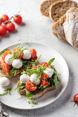 Sandwiches with tomatoes, mozzarella balls, arugula on white plate light textured background with ingredients close up. Italian bruschetta set, antipasti