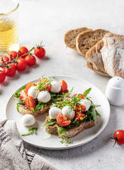 Sandwiches with tomatoes, mozzarella balls, arugula on white plate light textured background with ingredients. Italian bruschetta set, antipasti