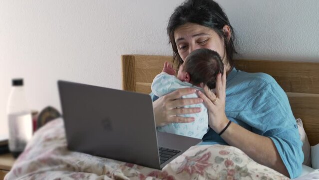 Mother smelling baby's diapers to check for poop while multitasking work and parenting in front of a computer, laid in bed, highlighting the balance of professional and personal life