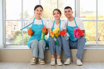 Team of young janitors with cleaning supplies sitting on windowsill in room