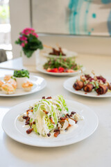 Wedge salad with bacon crumbles and white dressing on a white plate. Various other food on plates in background. Vertical color photo