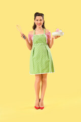 Portrait of beautiful pin-up woman in apron with whisk and sweets on yellow background