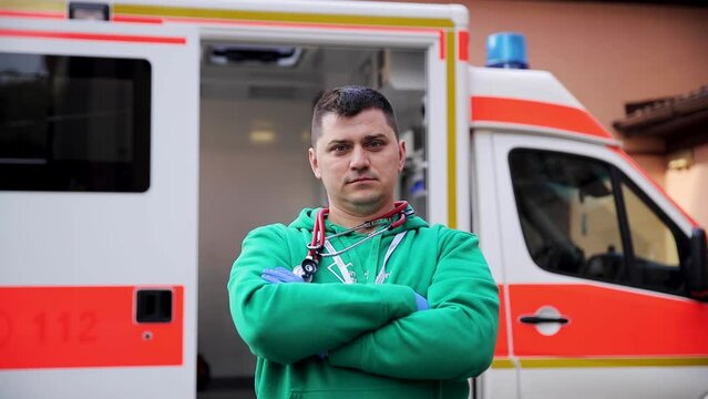 Caucasian male doctor with stethoscope on neck posing on camera with ambulance car on background. Competent paramedic wearing green uniform standing on city street