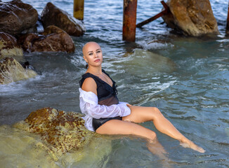 women with alopecia on beach opposing.beautiful female lady talking at smartphone sitting on lounge.girl in swimming suit and white shirt on rock, in water.sea,vacation background.