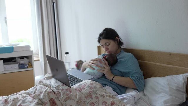 Mother cradling her newborn baby close to chest while balancing work life in front of laptop, laid in bed during infant's first week of life, capturing the essence of multitasking and dedication