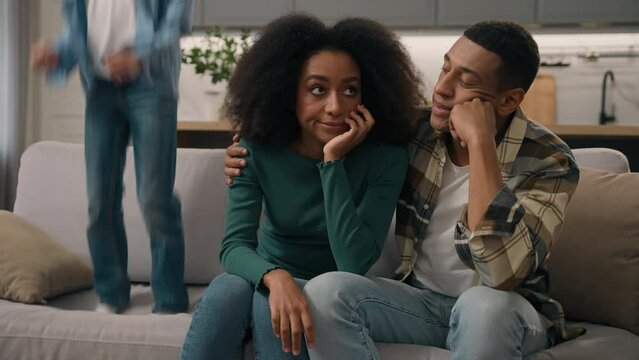 Stressed African American married couple tired mom dad sitting couch suffer naughty noisy shout little boy active son tantrum jump sofa house room. Unhappy female woman male man irritated by child kid