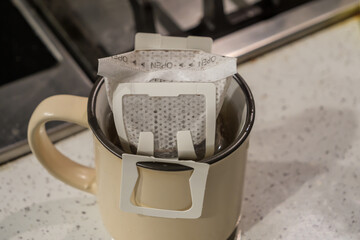 A drip filter coffee bag is set in a small ceramic mug, ready for brewing coffee. The mug is placed on a speckled kitchen countertop beside a stove.