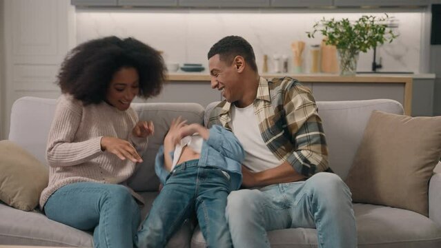 Playful parents mother mom female woman father dad male man tickling hug sincere laughing funny game ticklish having fun little boy child son kid living room bed. African American family cuddle indoor