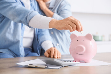 Mature couple putting coin into piggy bank in kitchen, closeup