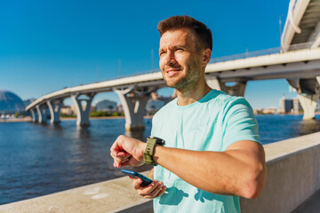 A man checks his watch while holding a phone, standing by a waterfront with a bridge and blue sky...