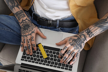Young tattooed man with disposable electronic cigarette and laptop sitting on armchair at home