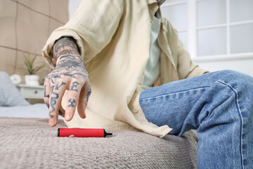 Young tattooed man sitting on bed and using disposable electronic cigarette at home