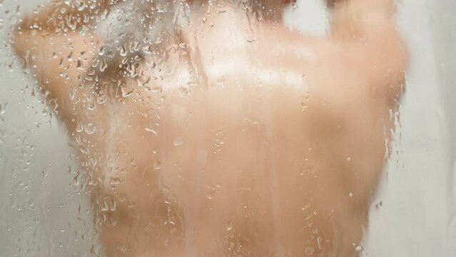 Blurred Silhouette of a Naked Woman in the Shower Cabin Washing Her Long Brown Hair, Water Droplets on the Glass.