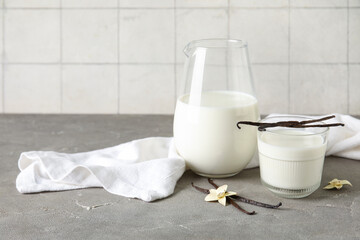 Glass and jug of fresh vanilla milk with sticks and flowers on grey table